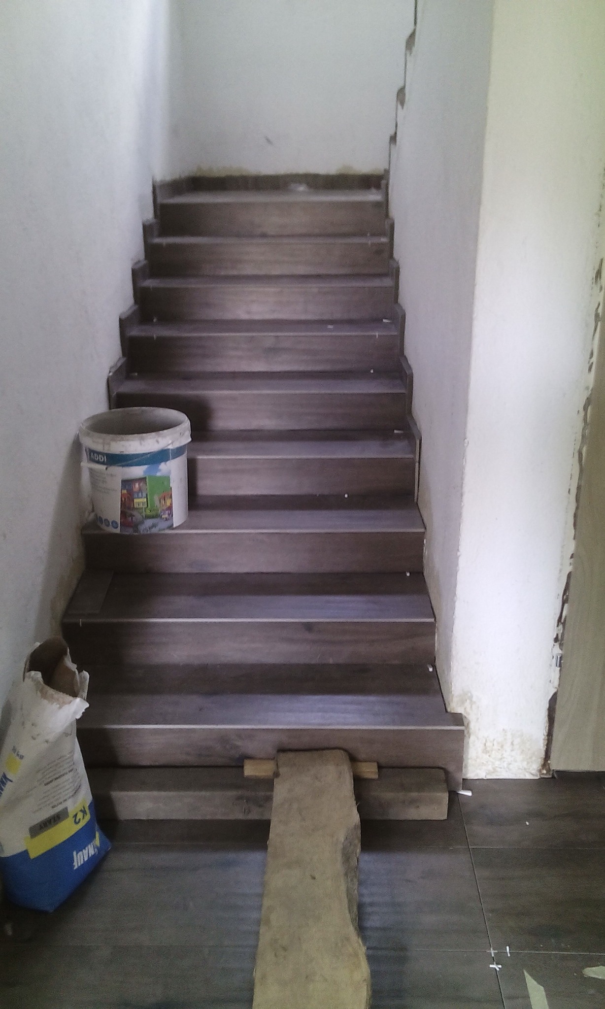 Schody wyłożone płytkami imitującymi drewno, z wiadrem farby i workiem zaprawy obok, na etapie wykończenia.