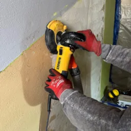 Szlifierka DeWalt z odkurzaczem w dłoniach w rękawicach Milwaukee szlifuje ścianę pokrytą gładzią, przygotowując ją do malowania.