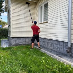 KAMAR - Mężczyzna w czerwonej koszulce czyści elewację domu sidingiem za pomocą szczotki na teleskopowym kiju; widoczne ślady czyszczenia i mokra powierzchnia.