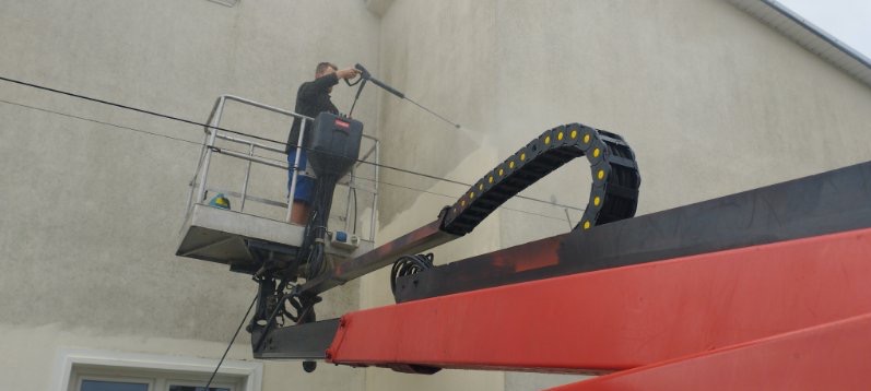 Pracownik na podnośniku koszowym czyści elewację budynku za pomocą myjki ciśnieniowej, widoczne różnice w czystości między umytą a brudną powierzchnią.