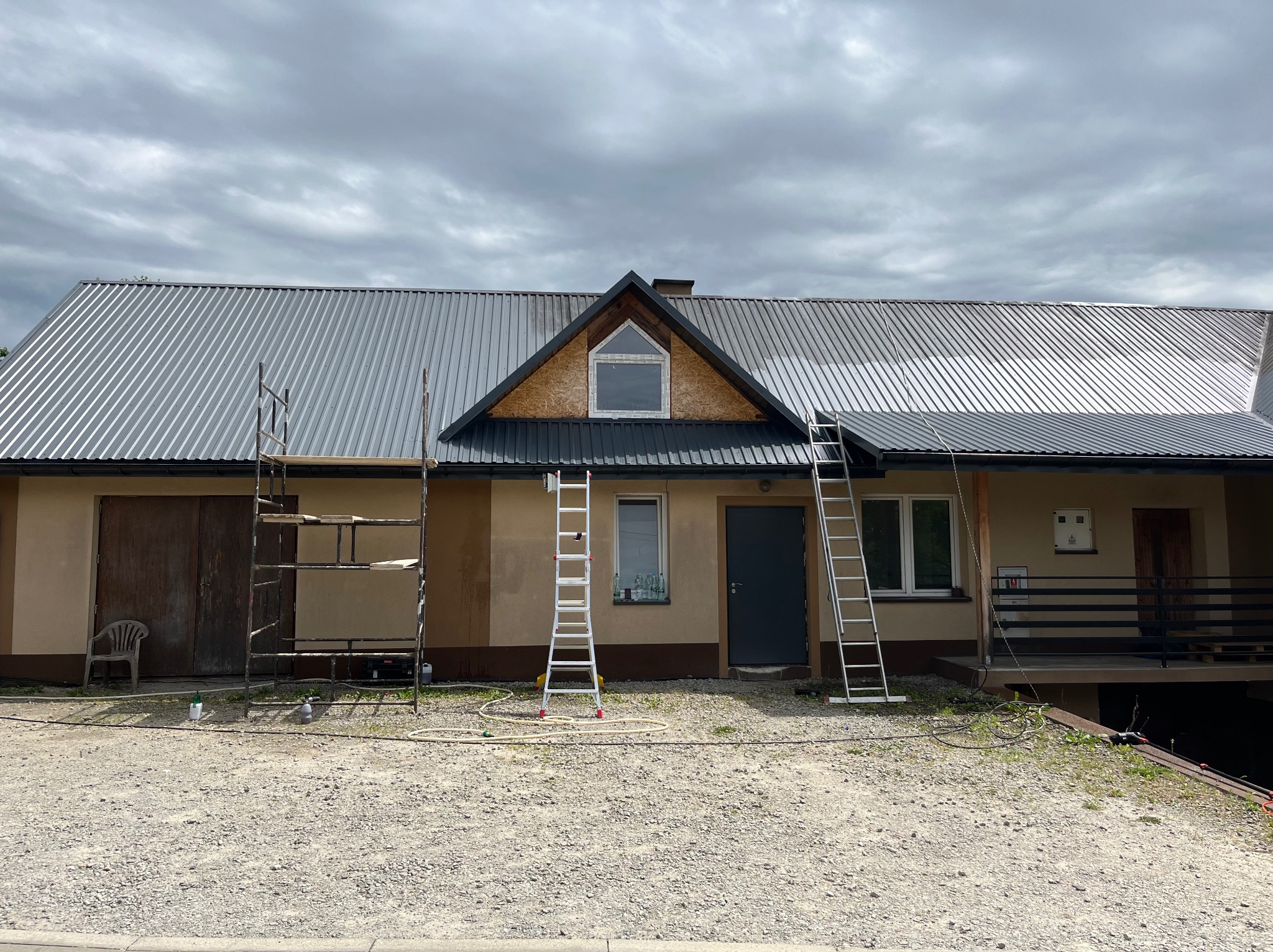 Dwukondygnacyjny budynek z blachy trapezowej na dachu w trakcie prac remontowych, widoczne rusztowania i drabiny opierające się o elewację.