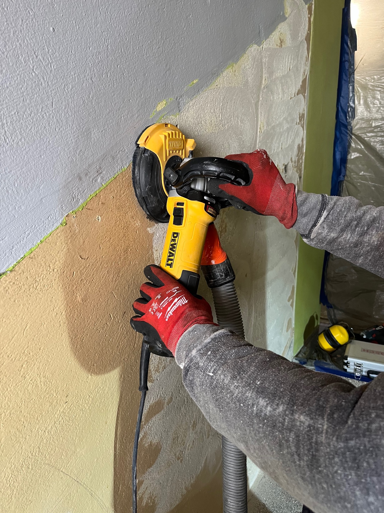 Szlifierka DeWalt z odkurzaczem w dłoniach w rękawicach Milwaukee szlifuje ścianę pokrytą gładzią, przygotowując ją do malowania.