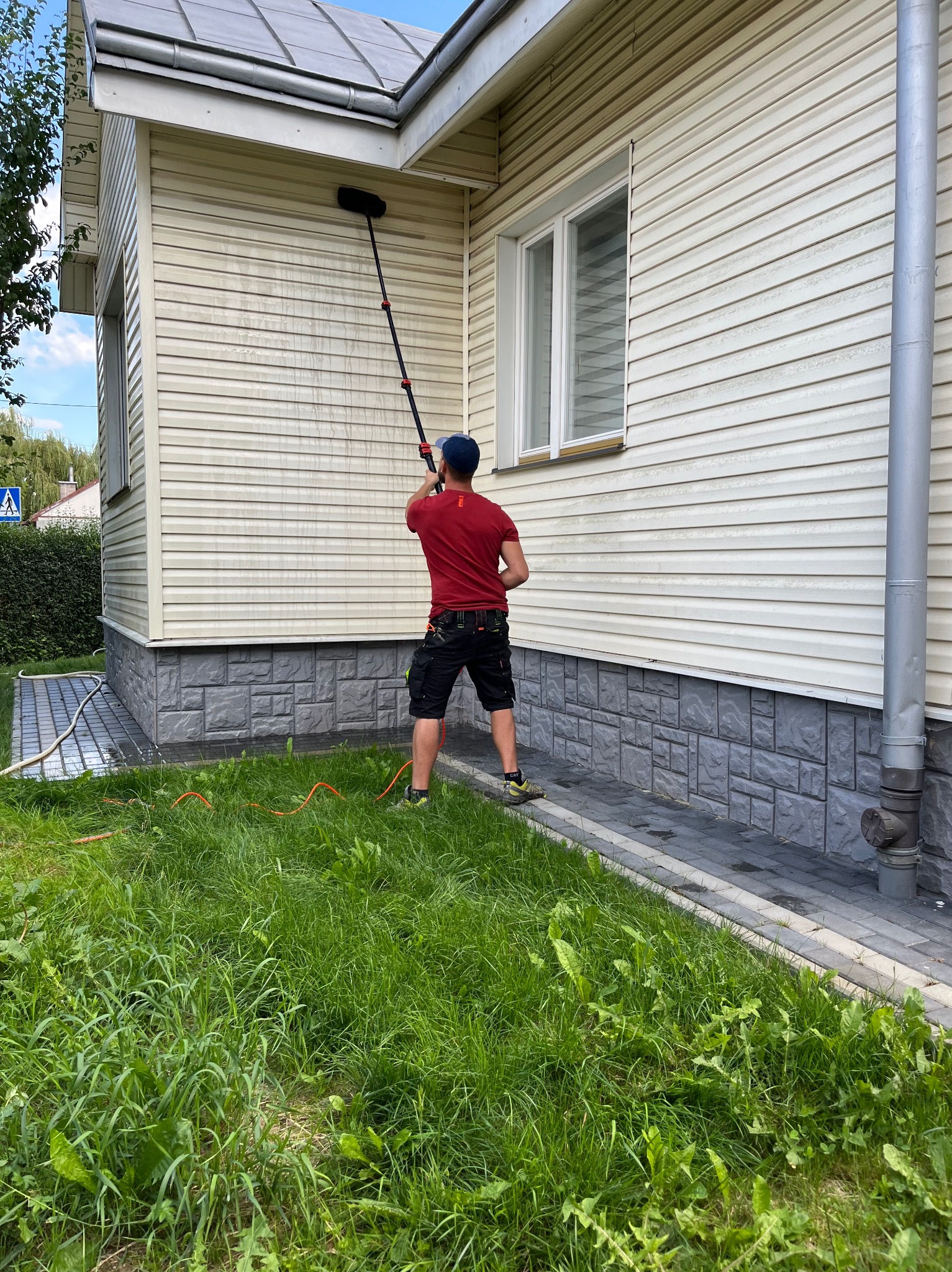 Mężczyzna w czerwonej koszulce czyści elewację domu sidingiem za pomocą szczotki na teleskopowym kiju; widoczne ślady czyszczenia i mokra powierzchnia.