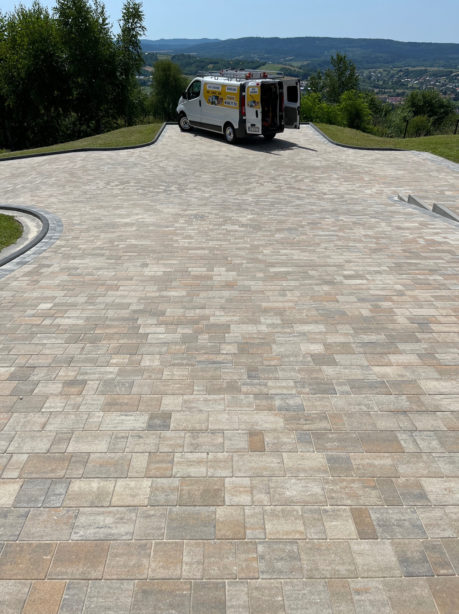 Ułożona kostka brukowa w odcieniach szarości i brązu na podjeździe z widocznym autem dostawczym firmy w tle i zielonym krajobrazem.