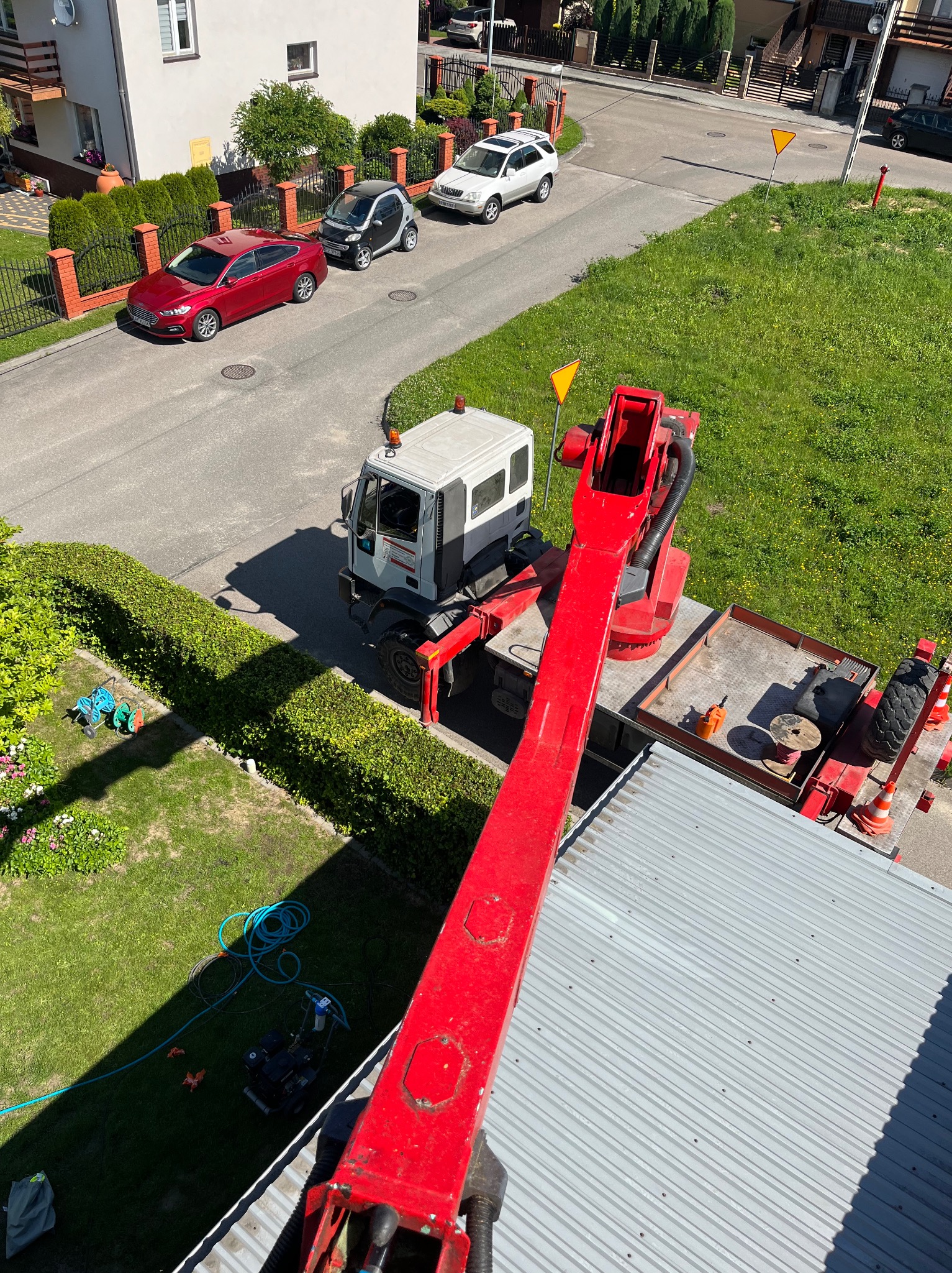 Widok z góry na podnośnik koszowy z wysięgnikiem nad szarym dachem blaszanym, w tle zabudowa jednorodzinna, trawniki i zaparkowane samochody.