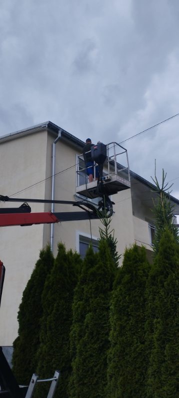 Pracownik na podnośniku koszowym przy elewacji dwupiętrowego domu z beżową fasadą, widoczne rynny i okno, na pierwszym planie wysokie tuje.