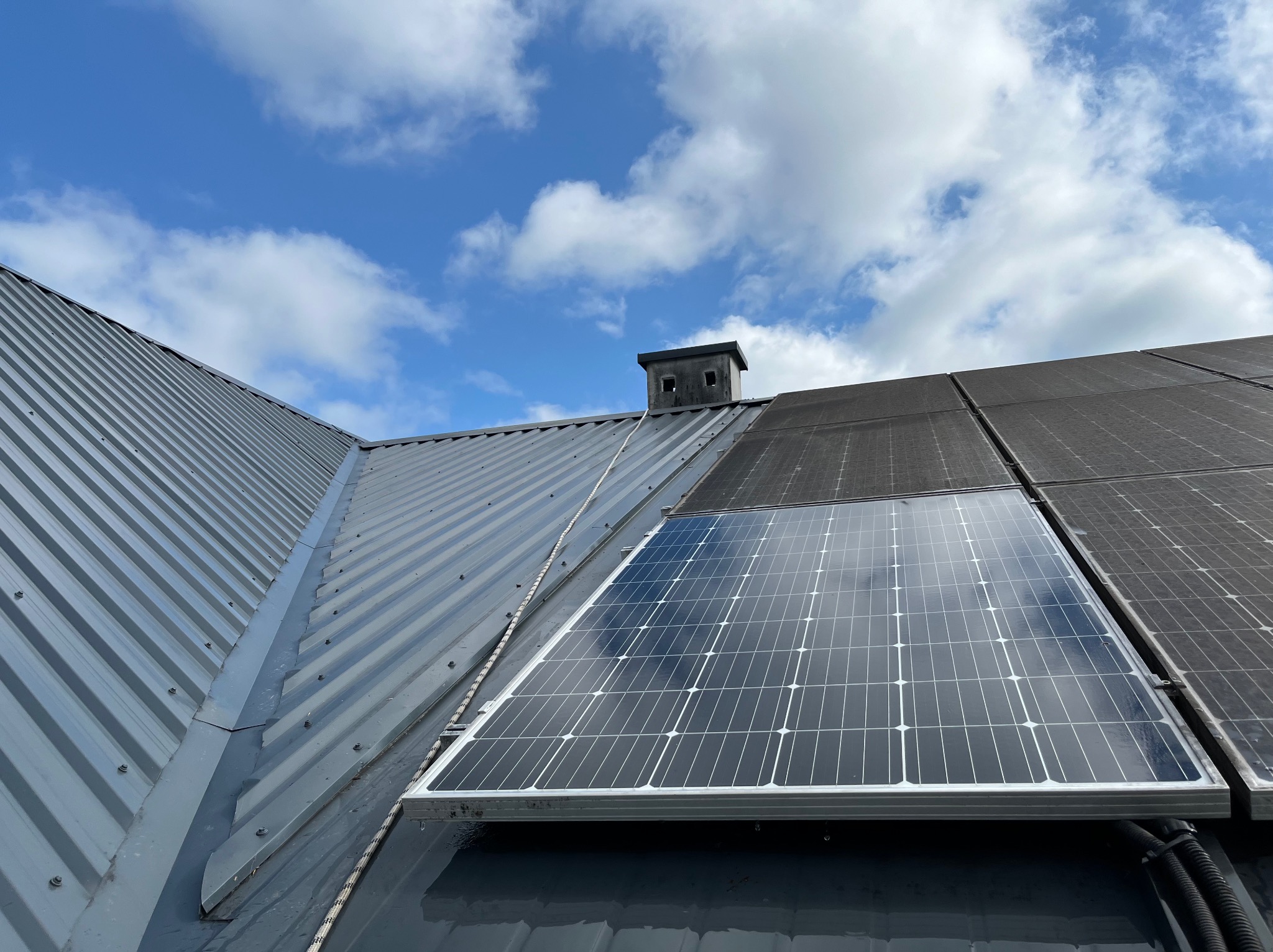 Panele słoneczne na metalowym dachu, jeden panel czysty, sąsiednie zabrudzone, komin w tle i błękitne niebo z chmurami.