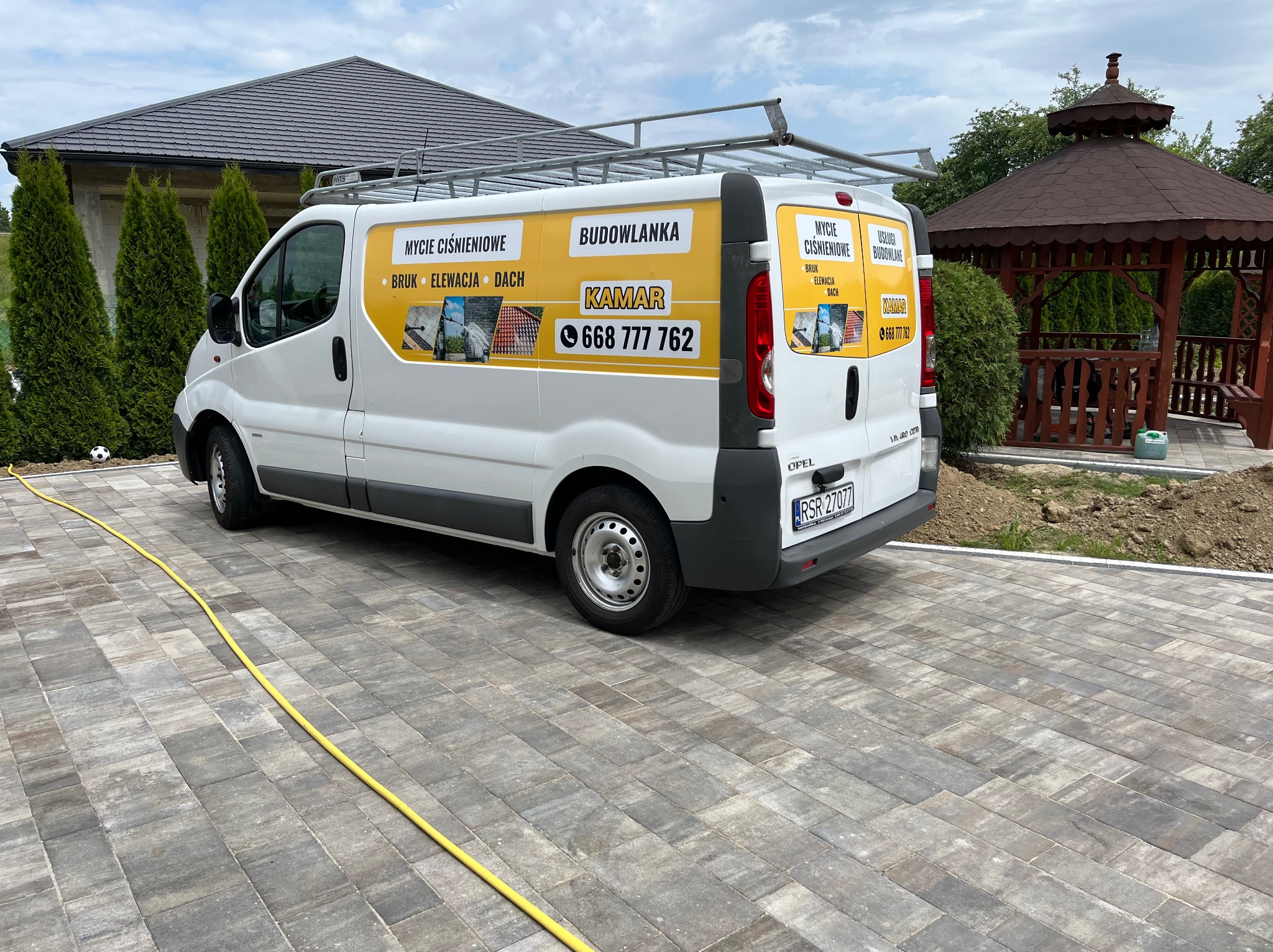 Biały samochód dostawczy firmy budowlanej Kamar z reklamą mycia ciśnieniowego bruku, elewacji i dachu, zaparkowany na kostce brukowej w pobliżu altany.