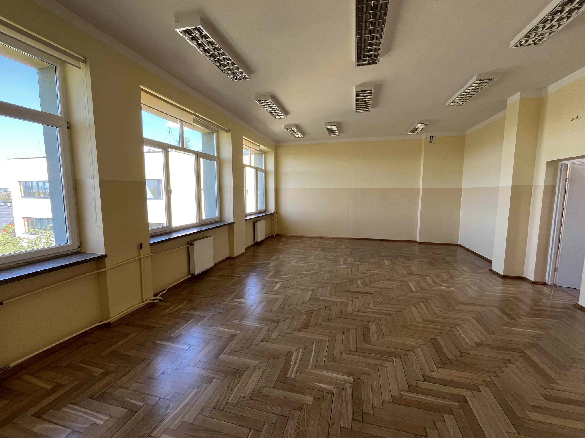 Jasne wnętrze sali z parkietem w jodełkę, rzędem okien i lampami sufitowymi. Ściany dwukolorowe, kremowo-żółte. Widok na odnowione pomieszczenie.