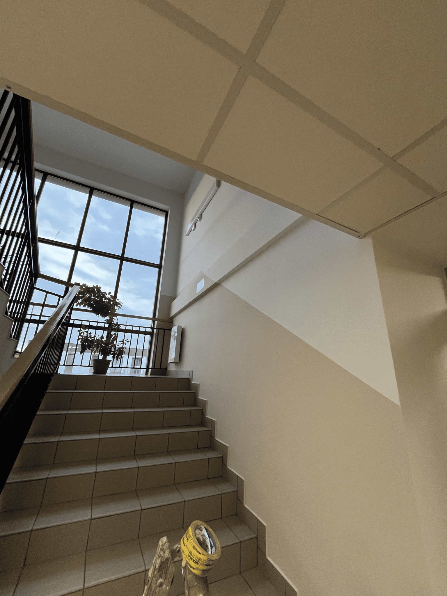 Klatka schodowa z oknem i rośliną w doniczce. Ściany pomalowane w dwóch odcieniach beżu. Na schodach leżą narzędzia malarskie.