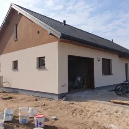 Dwukondygnacyjny dom z beżową elewacją i brązowym wykończeniem pod dachem, widoczne puste wiadra po farbie i rowery przed garażem.