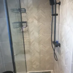 Nowoczesna kabina prysznicowa z czarną armaturą, półkami na kosmetyki i płytkami w jodełkę na ścianach.