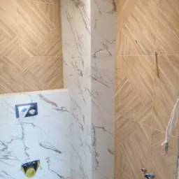 Ściana w łazience wyłożona płytkami imitującymi drewno w geometryczny wzór, kontrastująca z kolumną obłożoną białymi płytkami imitującymi marmur, widoczne instalacje wodne i fragment podtynkowego...