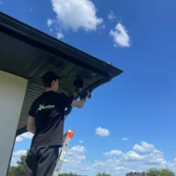 Mężczyzna na drabinie montuje czujnik pod dachem domu jednorodzinnego, na tle błękitnego nieba z chmurami. Widoczny wkrętak i logo firmy na koszulce.