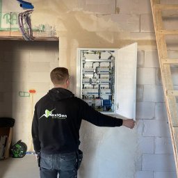 NextGen ElectroSystems Szymon Siepka - Elektryk w bluzie firmowej otwiera skrzynkę z bezpiecznikami na ścianie z betonu komórkowego. Widoczne kable i rury instalacyjne.