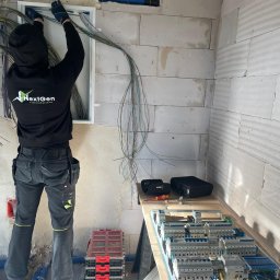 NextGen ElectroSystems Szymon Siepka - Elektryk w czarnej bluzie NextGen instaluje okablowanie w skrzynce elektrycznej na ścianie z betonu komórkowego. Widoczne narzędzia i rozdzielnica na stole.