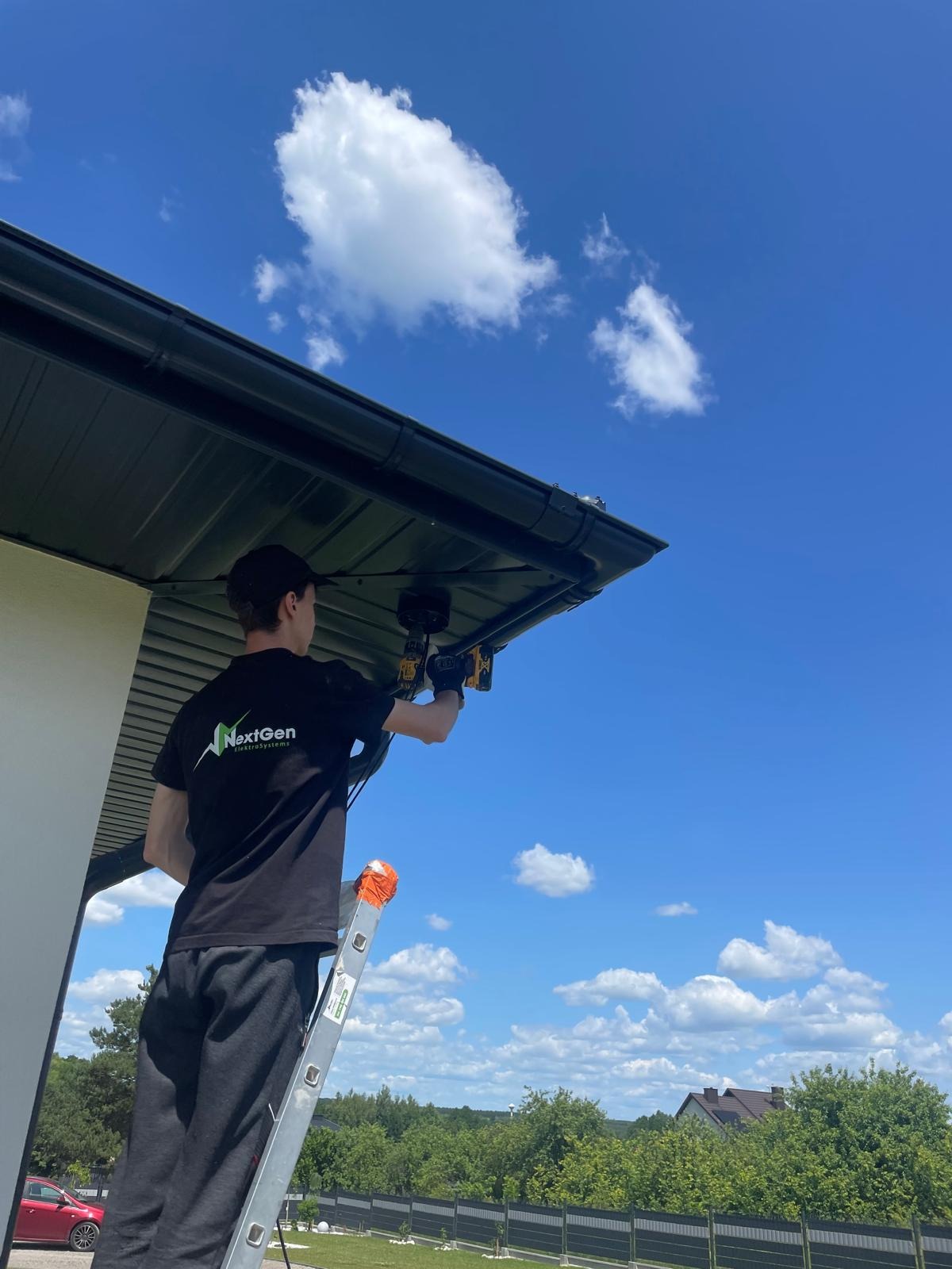 Mężczyzna na drabinie montuje czujnik pod dachem domu jednorodzinnego, na tle błękitnego nieba z chmurami. Widoczny wkrętak i logo firmy na koszulce.