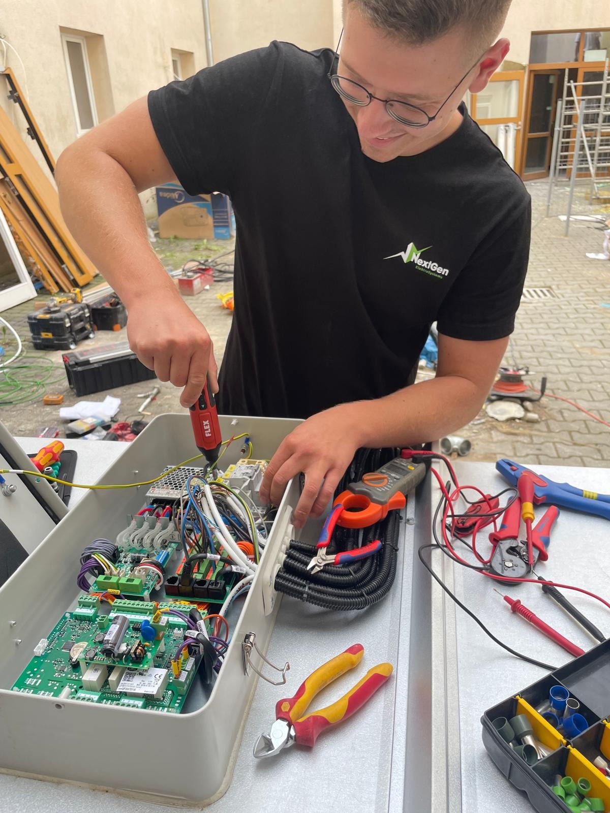 Elektryk w okularach, w czarnej koszulce z logo, skręca śrubokrętem elementy wewnątrz otwartej skrzynki z elektroniką. Obok leżą narzędzia.