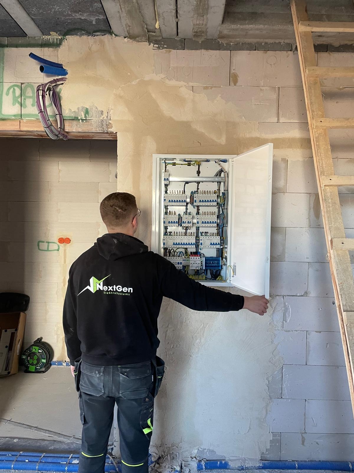 Elektryk w bluzie firmowej otwiera skrzynkę z bezpiecznikami na ścianie z betonu komórkowego. Widoczne kable i rury instalacyjne.