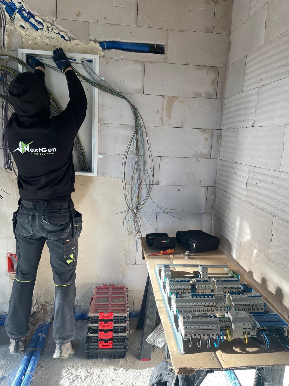 Elektryk w czarnej bluzie NextGen instaluje okablowanie w skrzynce elektrycznej na ścianie z betonu komórkowego. Widoczne narzędzia i rozdzielnica na stole.