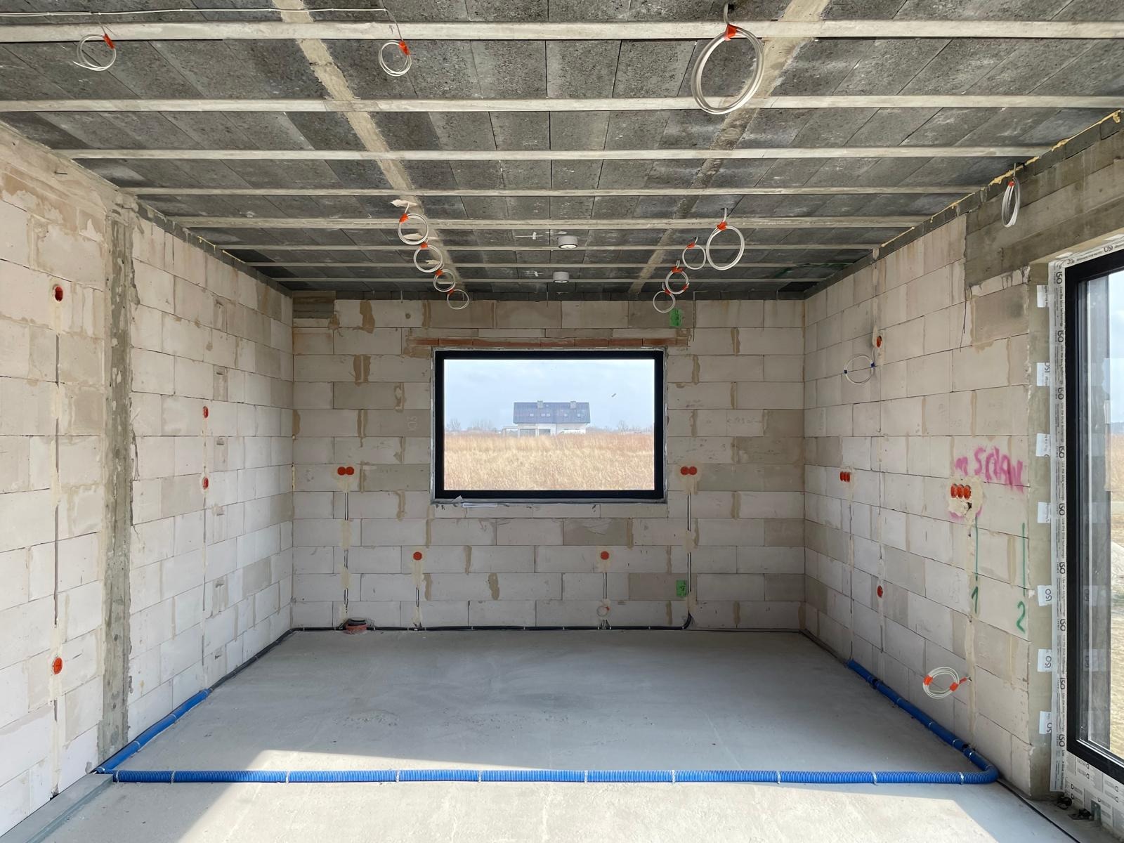 Surowy stan wnętrza domu w budowie. Widoczne instalacje w ścianach z betonu komórkowego i pod sufitem. W oknie widok na krajobraz.