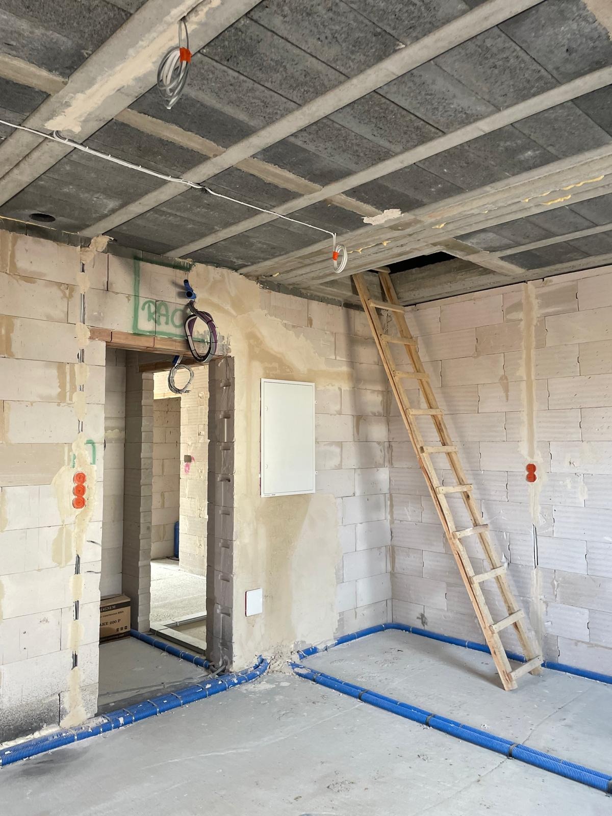 Surowy stan: instalacje elektryczne i rury w ścianach z betonu komórkowego, drabina drewniana prowadząca do otworu w stropie. W tle widoczne kable i puszki.