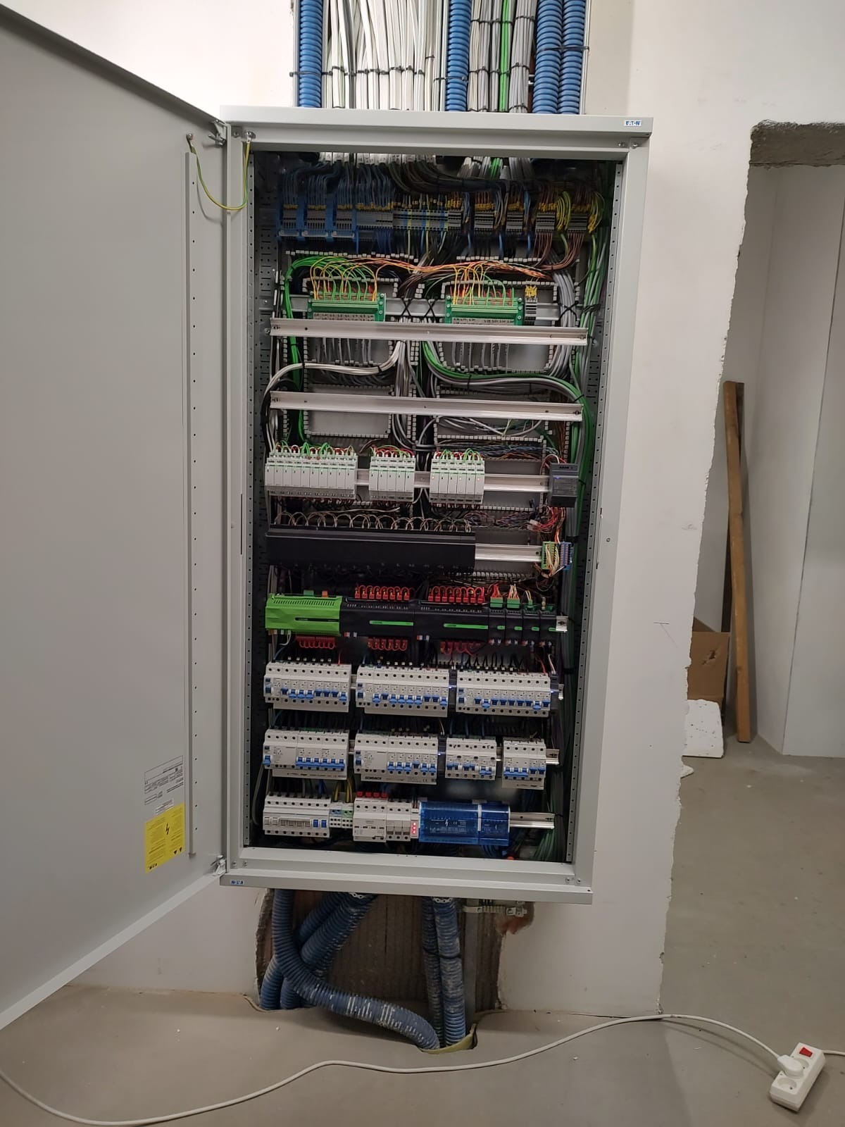 Otwarta szafa elektryczna z widocznymi bezpiecznikami, przewodami i elementami sterującymi, w surowym wnętrzu z widocznymi rurami instalacyjnymi.