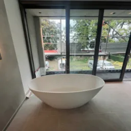 Nowoczesna, wolnostojąca wanna owalna w łazience z dużymi oknami przesuwnymi i widokiem na rusztowania oraz samochody dostawcze na zewnątrz.