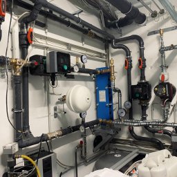 Andrzej Gmachowski - Rozbudowana instalacja hydrauliczna z czarnymi rurami PCV, pompami obiegowymi Grundfos, wymiennikiem ciepła i zbiornikiem wyrównawczym, widoczne manometry i zawory kulowe z pomarańczowymi dźwigniami.