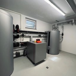 Andrzej Gmachowski - Nowoczesna kotłownia z szarymi zbiornikami i rurami, widoczne okno w tle, pomarańczowe narzędzia na szafce, minimalistyczny design.