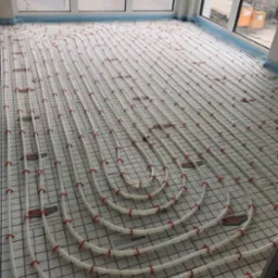 Białe rury ogrzewania podłogowego, mocowane czerwonymi klipsami do siatki na podłodze w jasnym pomieszczeniu z oknami, widoczne oznaczenia producenta na siatce.