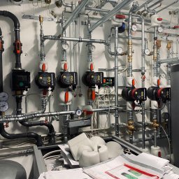 Andrzej Gmachowski - Rozbudowana instalacja grzewcza z wieloma pompami obiegowymi Grundfos, rurami i zaworami, widoczne manometry i izolacja termiczna, dokumentacja techniczna na pierwszym planie.