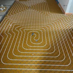 Instalacja ogrzewania podłogowego z białych rurek na brązowej izolacji podłogowej, układanych w pętle na dużej powierzchni pomieszczenia w trakcie remontu.