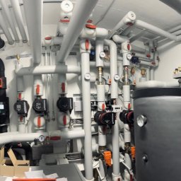 Andrzej Gmachowski - Rozbudowana kotłownia z licznymi rurami, zaworami, pompami obiegowymi i zbiornikami akumulacyjnymi, widoczne także elementy automatyki i instalacji elektrycznej.