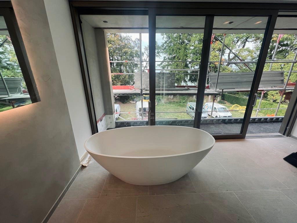 Nowoczesna, wolnostojąca wanna owalna w łazience z dużymi oknami przesuwnymi i widokiem na rusztowania oraz samochody dostawcze na zewnątrz.