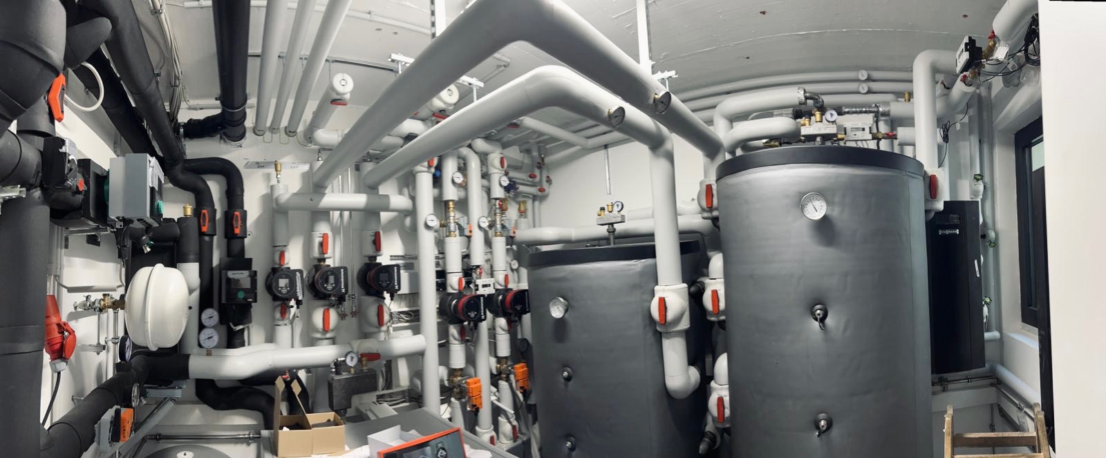 Skomplikowana instalacja grzewcza w kotłowni, widoczne dwa szare zbiorniki akumulacyjne z manometrami, liczne białe i czarne rury, pompy obiegowe i armatura, częściowo widoczna drabina i karton...