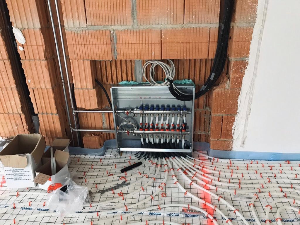 Rozdzielacz ogrzewania podłogowego zamontowany w surowym stanie, widoczne rury doprowadzające i odprowadzające wodę, instalacja na podłodze z siatką montażową, ściany w stanie deweloperskim z cegły...