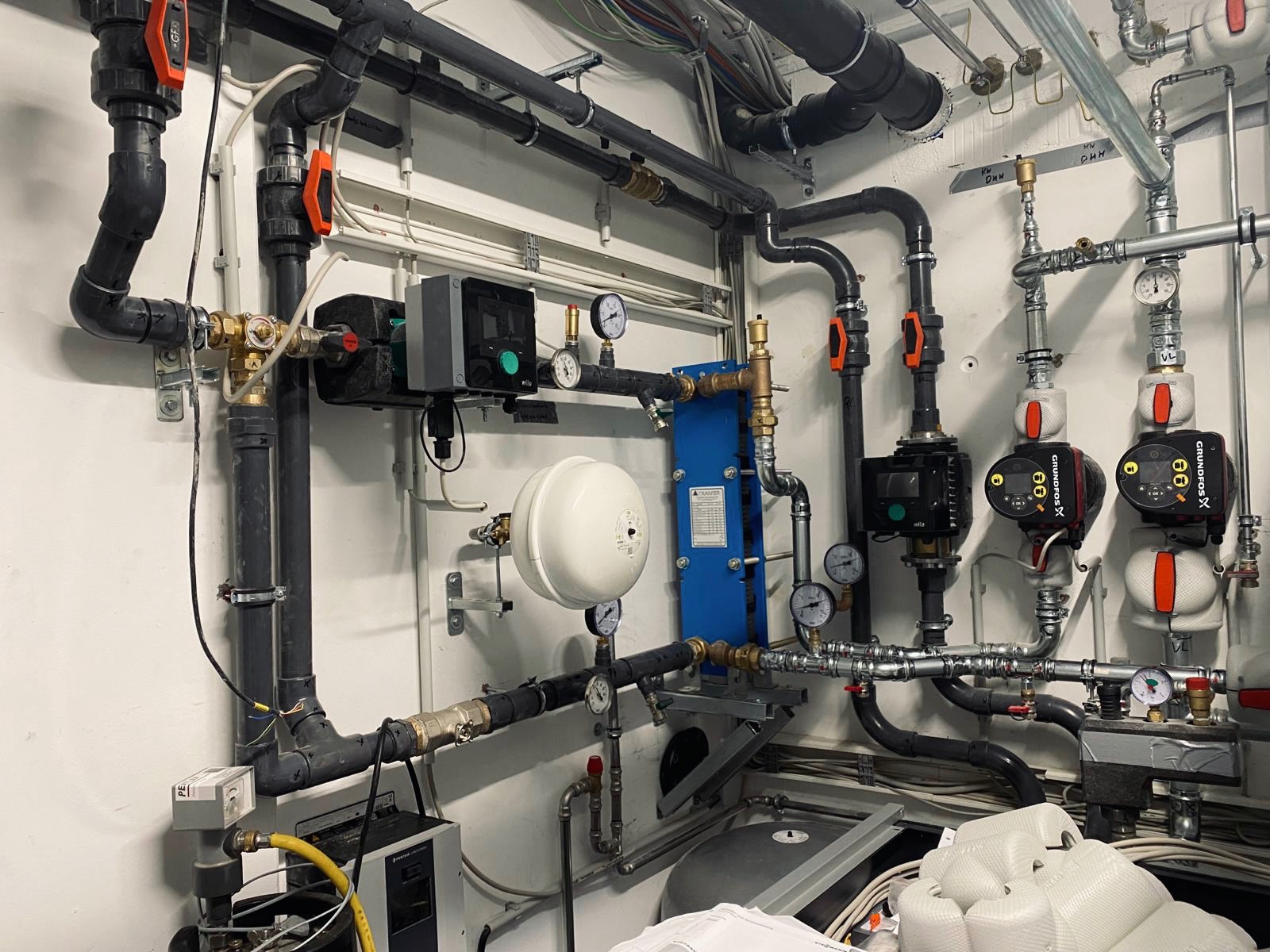 Rozbudowana instalacja hydrauliczna z czarnymi rurami PCV, pompami obiegowymi Grundfos, wymiennikiem ciepła i zbiornikiem wyrównawczym, widoczne manometry i zawory kulowe z pomarańczowymi dźwigniami.