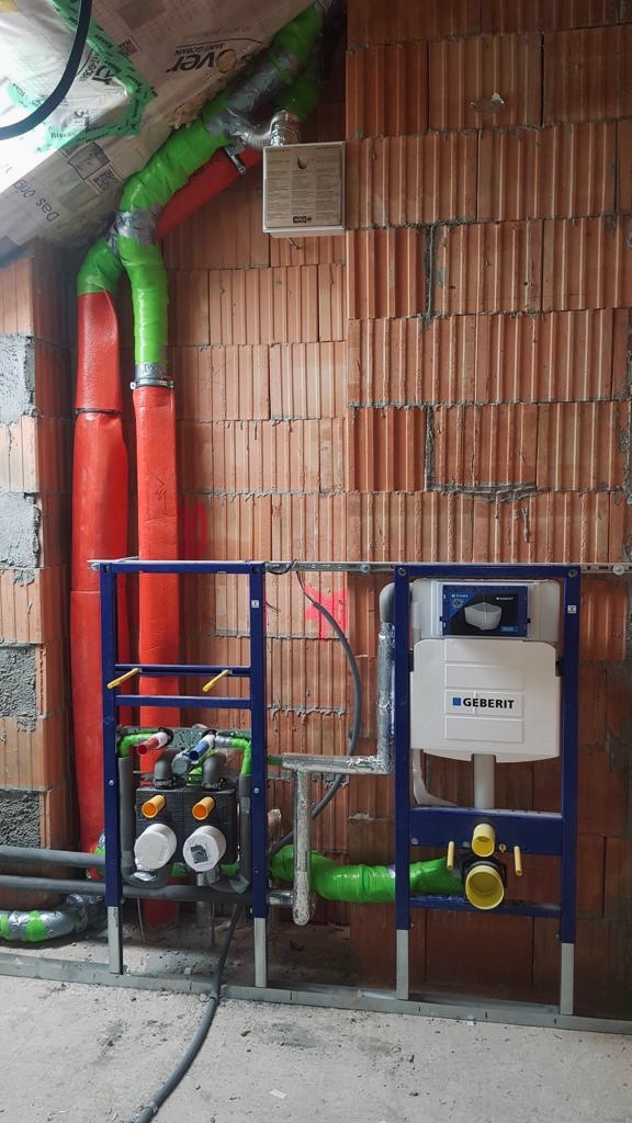 Instalacja podtynkowa Geberit Duofix z widocznymi rurami w izolacji (czerwonej i zielonej) oraz fragmentem ściany z cegły kratówki w tle. Na ścianie widoczna tabliczka informacyjna.