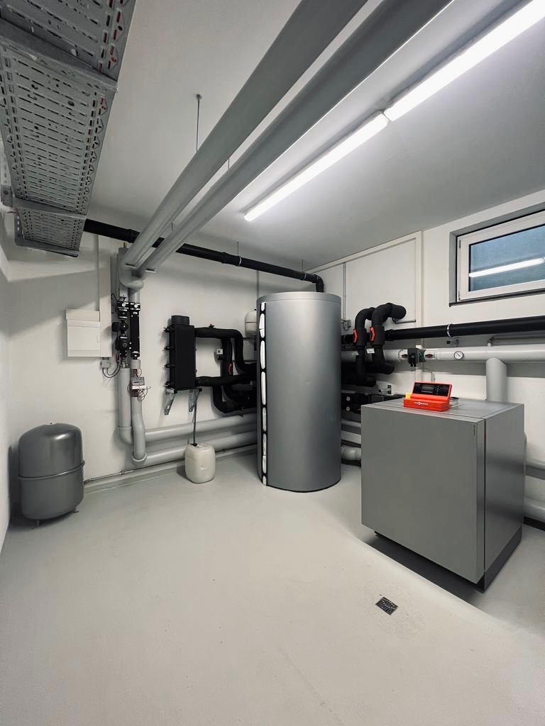 Szare pomieszczenie techniczne z zainstalowanym zbiornikiem akumulacyjnym, piecem, orurowaniem i elementami sterującymi, widoczne kanały kablowe pod sufitem.