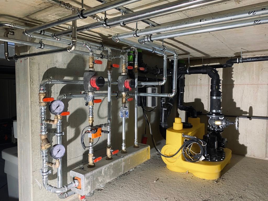 Kompleksowa instalacja rur stalowych i plastikowych z manometrami, zaworami, pompami obiegowymi i żółtą przepompownią ścieków w piwnicy z surowym betonowym sufitem.