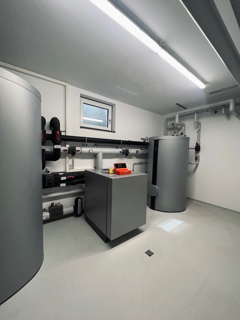 Nowoczesna kotłownia z szarymi zbiornikami i rurami, widoczne okno w tle, pomarańczowe narzędzia na szafce, minimalistyczny design.