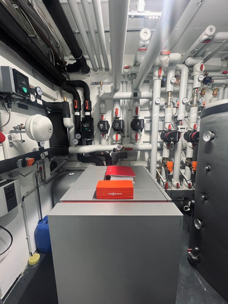 Pomieszczenie kotłowni z piecem gazowym Viessmann, widoczne liczne rury, zawory i manometry, na piecu leżą dokumenty i pomarańczowe pudełko.