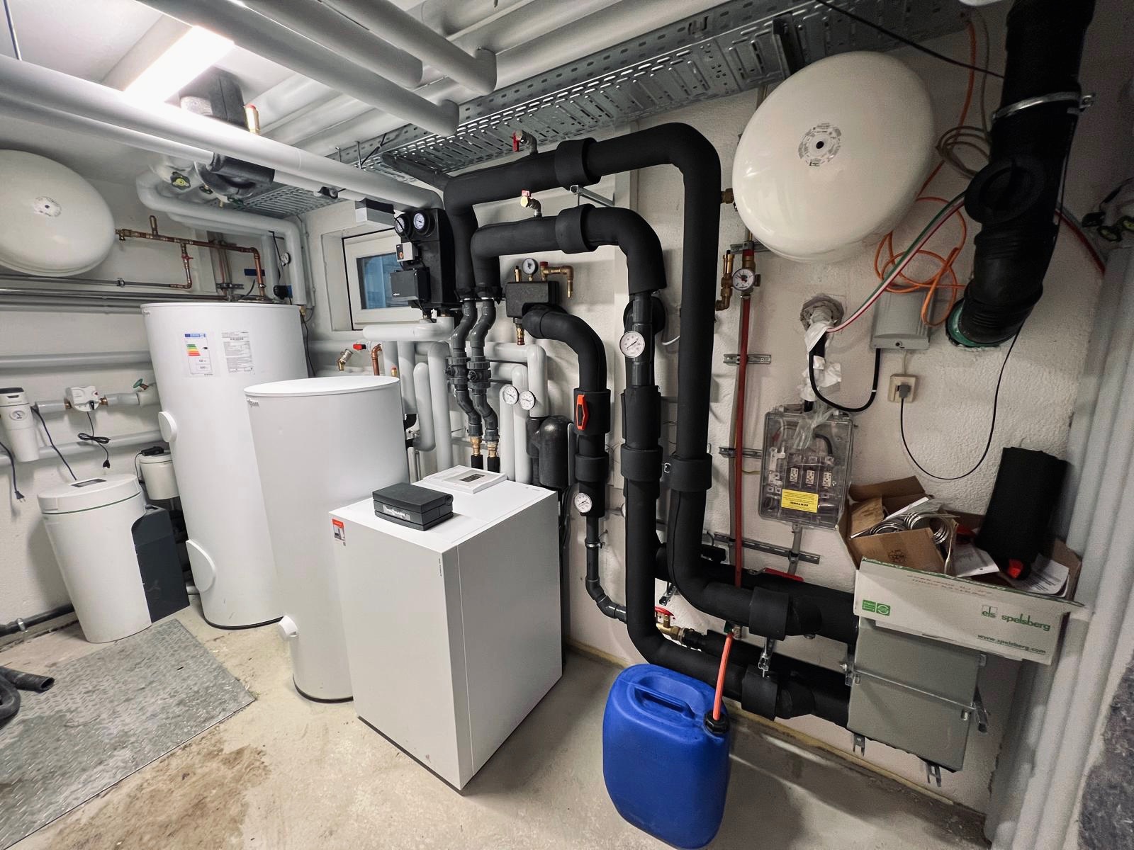 Kotłownia z białymi zbiornikami, czarnymi rurami izolowanymi, niebieskim kanistrem i skrzynką elektryczną na ścianie, widok na pomieszczenie technicznym.