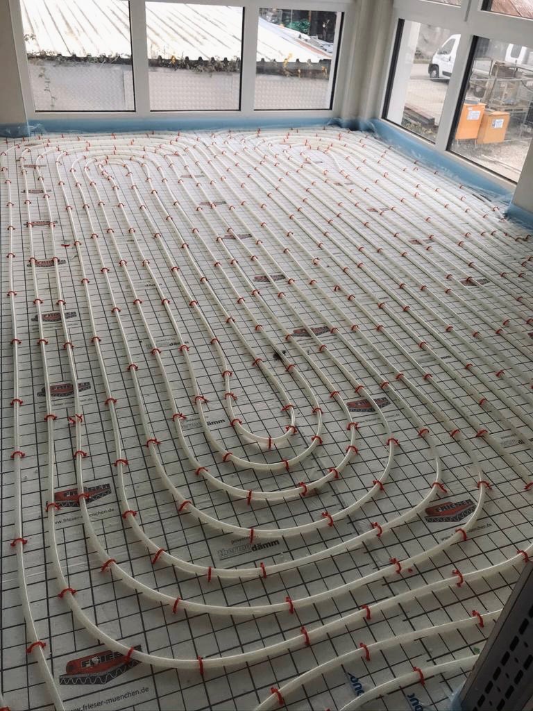 Białe rury ogrzewania podłogowego, mocowane czerwonymi klipsami do siatki na podłodze w jasnym pomieszczeniu z oknami, widoczne oznaczenia producenta na siatce.