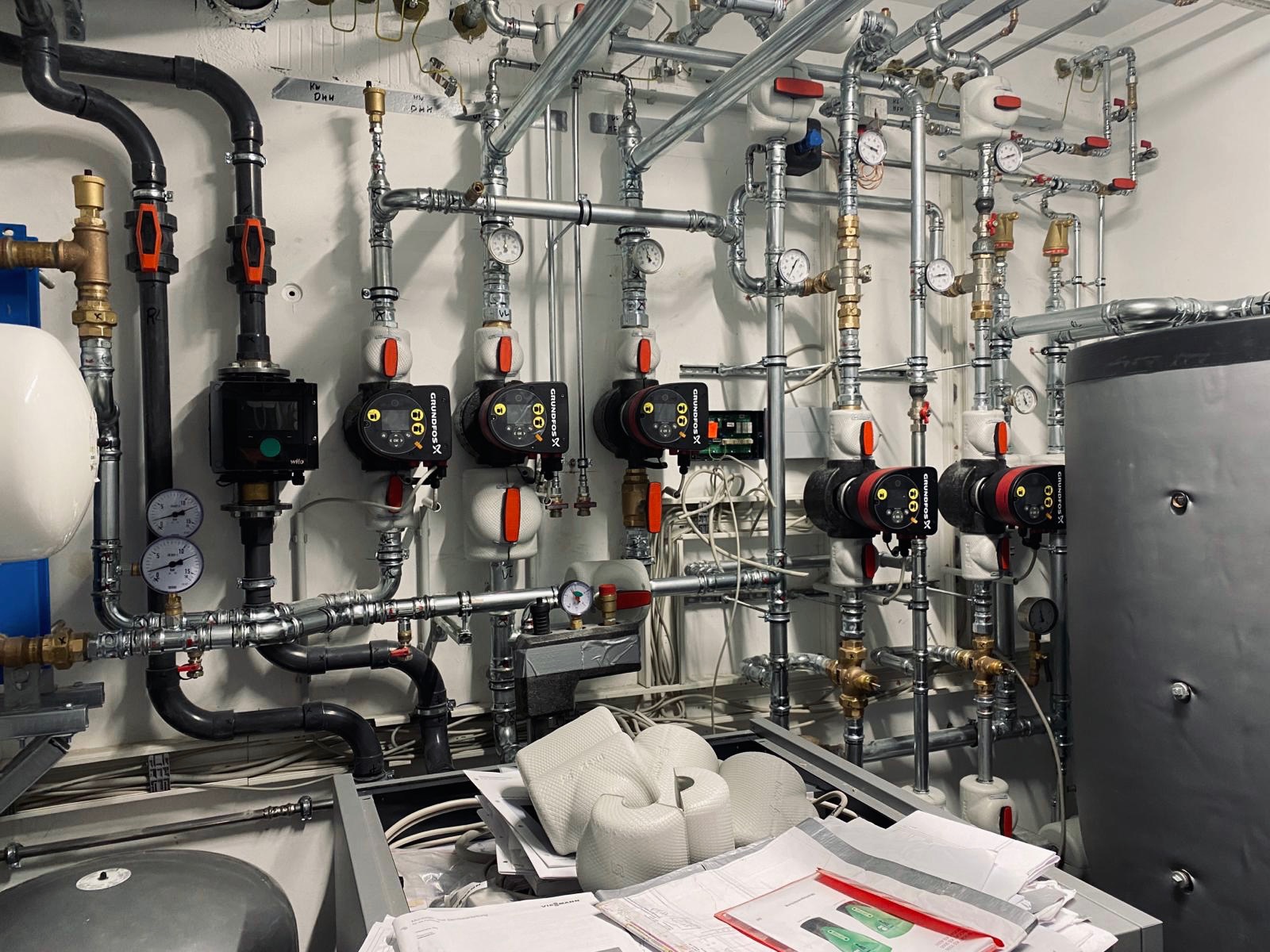 Rozbudowana instalacja grzewcza z wieloma pompami obiegowymi Grundfos, rurami i zaworami, widoczne manometry i izolacja termiczna, dokumentacja techniczna na pierwszym planie.