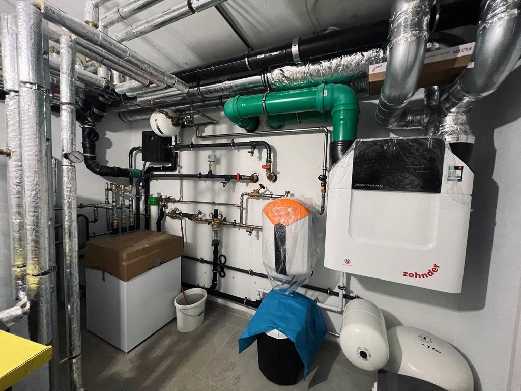 Węzeł cieplny w piwnicy: biały piec Zehnder, rury instalacji grzewczej z izolacją, zielone rury wentylacyjne, zbiorniki wyrównawcze, manometry i zawory.