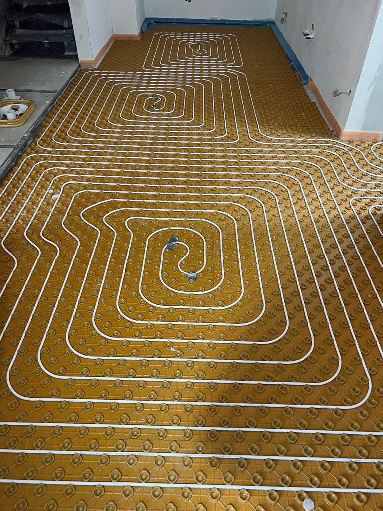 Instalacja ogrzewania podłogowego z białych rurek na brązowej izolacji podłogowej, układanych w pętle na dużej powierzchni pomieszczenia w trakcie remontu.