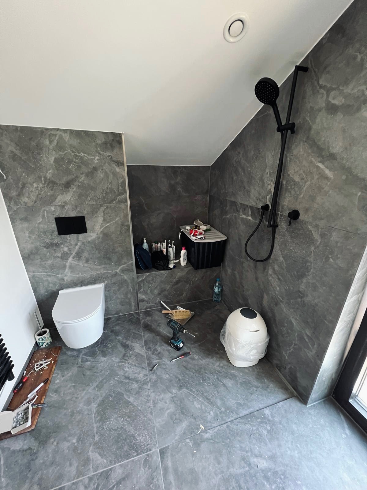 Łazienka w trakcie wykończenia: szare marmurowe płytki, biała toaleta podtynkowa, czarna deszczownica, narzędzia i akcesoria budowlane na podłodze.