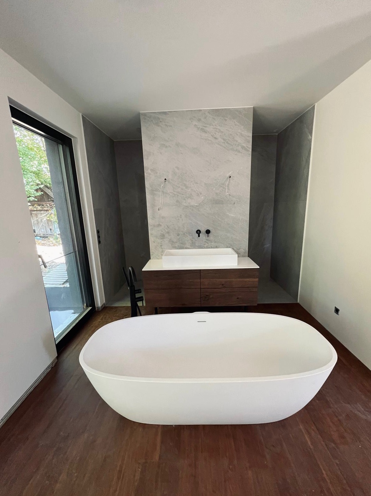 Nowoczesna łazienka w trakcie wykańczania: wolnostojąca biała wanna owalna, szafka pod umywalkę z drewnianymi frontami, marmurowe płytki ścienne, widoczne przewody instalacji przed podłączeniem...
